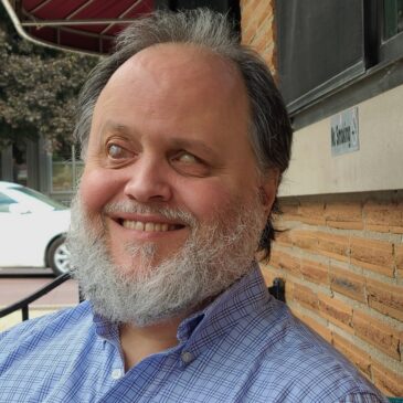 A man of European descent is smiling at us and at the sunny summer day around him. His grey beard might suggest a calm and wise demeanor, but those eye-crinkles are all mischief, waiting to drop double and triple entendres on the unwary.