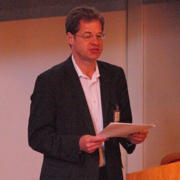 A man of European decent is delivering a talk with a sheaf of notes in his hand. He's ignoring the notes, however, and he has wandered away from the podium. He leans in toward the audience with a look of excitement such as he probably had when he was discovering a new cool book at the library as a small boy.