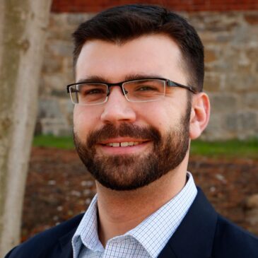 Dr. Liam Daley sports a trim beard and glasses to go with his big smile. He's somewhere outdoors with a stone wall in the background. He's clearly got laugh crinkles by his eyes.