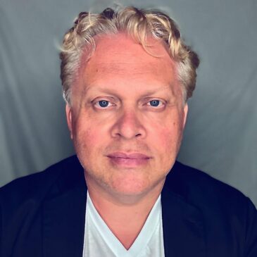 Tony is giving us a frank, direct stare, blond curls,a nd a hint of a grin. He is a man of European ancestry, dressed in a white v-necked shirt and blue jacket.