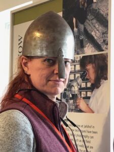 Grace Alden is a redheaded woman of European ancestroy who really looks good in an iron age Pictish helmet with one of those nose-guard things. She is standing in the visitor center of Jarlsberg, an archaeological site in Shetland, just bonding with the ancestors