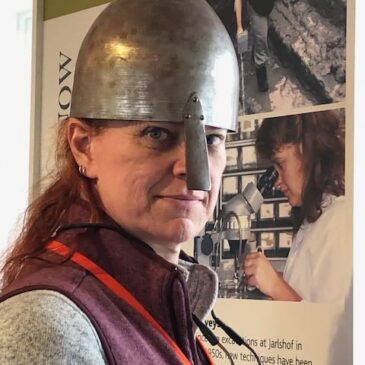 Grace Alden is a redheaded woman of European ancestroy who really looks good in an iron age Pictish helmet with one of those nose-guard things. She is standing in the visitor center of Jarlsberg, an archaeological site in Shetland, just bonding with the ancestors