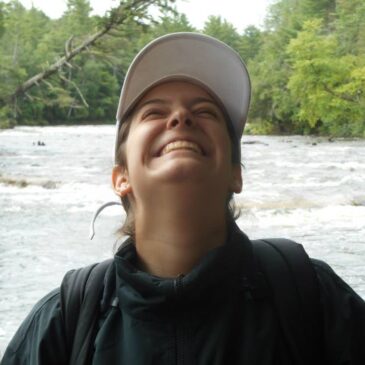 Cate is outdoors where she's happiest— and I have honestly never seen a bigger smile, ear-to-ear and apple-cheeked. Whitewater right behind her suggests that this photo was taken when she was in a kayak. She's a young woman of European descent with brown hair probably in a ponytail wearing a hoodie and a ball cap to prevent sunburn on this gorgeous, sunny day. Her face is turned upward toward the canopy of nearby trees as though she is greeting all of her squirrel friends.
