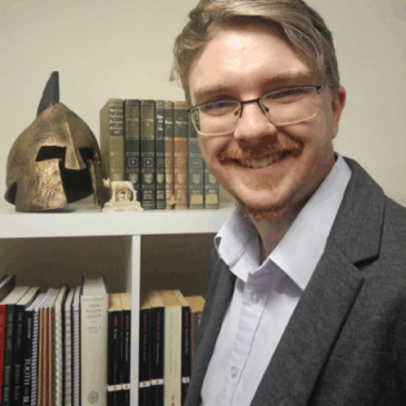 Julian Barr's bespectacled smile reveals a dimple or two in his neatly-trimmed red-blond goatee. He wears a crisp blue shirt and grey jacket and stands partly turned toward his collection of books—young paperbacks and venerable old hardbacks—which are punctuated by a wee replica statue of Lupa suckling Romulus and Remus and a replica bronze helm which looks like the one on the cover of the Chickering Beowulf, but given Dr. Barr's interest in rem Romanorum, it might be Roman, so I'll have to ask him about that