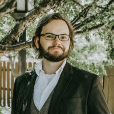 Patrick Lyon cuts a very dapper Hobbitty vibe in patterned waistcoat, green and blue kilt pleated to sett, kilt belt and brown leather sporran. His brown beard and horn rimmed glasses furthers the impression, perhaps as the head of school in Hobbiton or the first Hobbit professor of poetry at the University of Minas Tirith. He's surrounded by a lovely lush garden and wooden fences, and he's got a very kind smile.