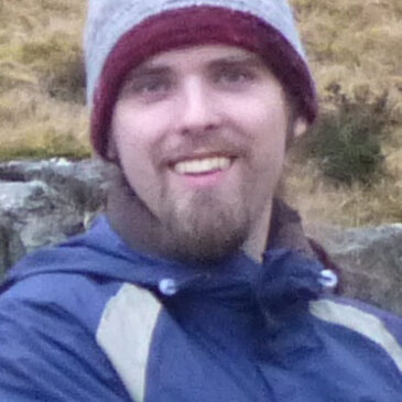 Nelson Goering is probably hiking as this photo is being shot. He's got an excited, clear-eyed expression, he's wearing a warm hat and a blue parka, and we see scrub and rocks on a slope behind him. He looks like he's having the best time ever!