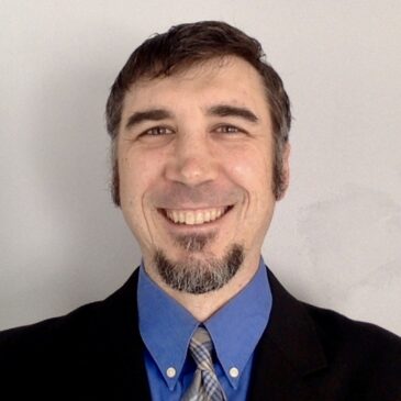 Joel has a broad smile and twinkling eyes above a salt-and-papper goatee. He is a man of apparently European ancestry wearing jacket, blue collared shirt and a stripey necktie tied in a half-Windsor.