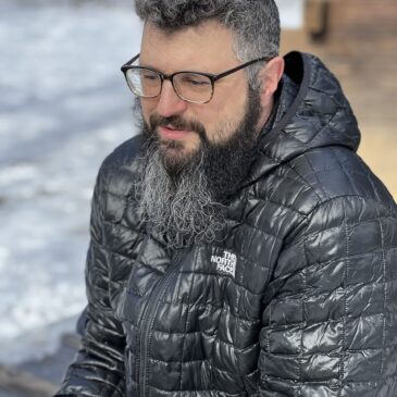 Shawn Gaffney, a scholar with salt-and-pepper hair and long beard sits outdoors wearing horn-rimmed glasses, a puffy dark jacket, and a faraway gaze.