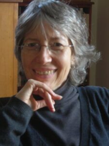 A graceful woman with salt-and-pepper hair, a broad smile, and a charcoal grey turtleneck and sweater rests her chin on her hand.