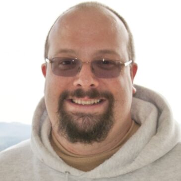 Trevor has such a warm smile! His brown goatee, slightly darkened glasses, and very short hair are the dark points in this outdoor photo of Trevor in a comfy grey hoodie.