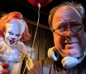 A gentleman of European extraction wearing headphones around his neck is looking over his spectacles at a truly terrifying clown doll. there's a bloon. I can't even keep alt-texting, the clown is so disturbing.